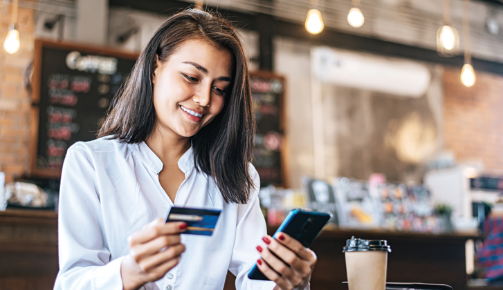 Young woman at a cofee shop holds her phone in one hand and her financial card in another.