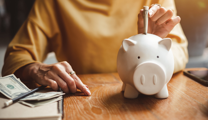 A woman sits at a wooden table with dillar bills near her left hand, depositing one into a white piggy bank.