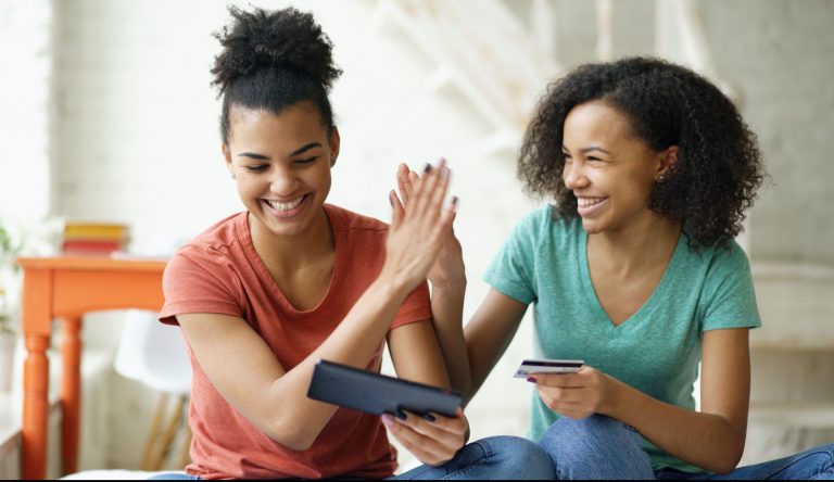 Two cheerful mixed race curly girlfriends shopping online with tablet computer and credit card at home